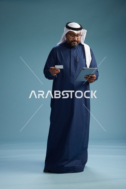 رجل سعودي يرتدي ثوب كحلي وغتره بيضاء يستخدم جهاز التابليت, الجسم بالطول الكامل, خلفيه زرقاء