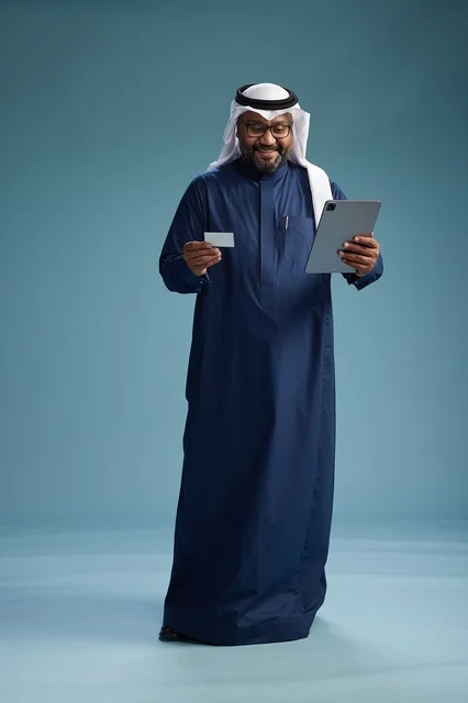 A Saudi man wearing a navy blue thobe and a white ghutrah is using a tablet, full-body shot, blue background. A Saudi man wearing a navy blue thobe and a white ghutrah is using a tablet, full-body shot, blue background.