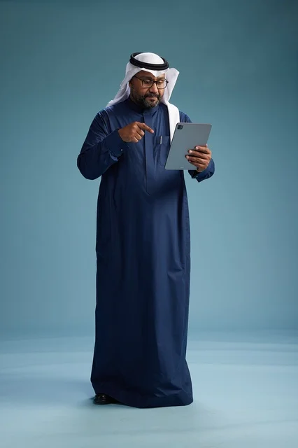 A Saudi man wearing a navy blue thobe and a white ghutrah is using a tablet, full-body shot, blue background.
