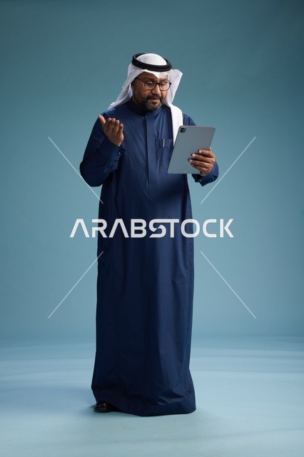 رجل سعودي يرتدي ثوب كحلي وغتره بيضاء يستخدم جهاز التابليت, الجسم بالطول الكامل, خلفيه زرقاء