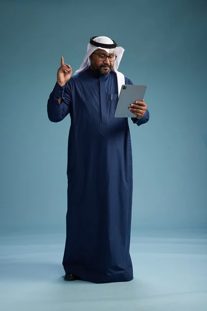 رجل سعودي يرتدي ثوب كحلي وغتره بيضاء يستخدم جهاز التابليت, الجسم بالطول الكامل, خلفيه زرقاء