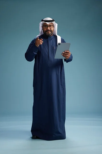 A Saudi man wearing a navy blue thobe and a white ghutrah is using a tablet, full-body shot, blue background.