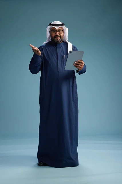 رجل سعودي يرتدي ثوب كحلي وغتره بيضاء يستخدم جهاز التابليت, الجسم بالطول الكامل, خلفيه زرقاء