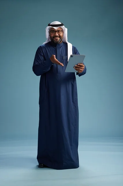 رجل سعودي يرتدي ثوب كحلي وغتره بيضاء يستخدم جهاز التابليت, الجسم بالطول الكامل, خلفيه زرقاء
