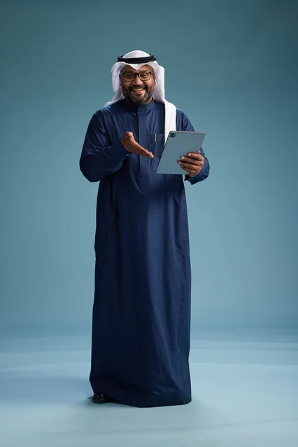 رجل سعودي يرتدي ثوب كحلي وغتره بيضاء يستخدم جهاز التابليت, الجسم بالطول الكامل, خلفيه زرقاء