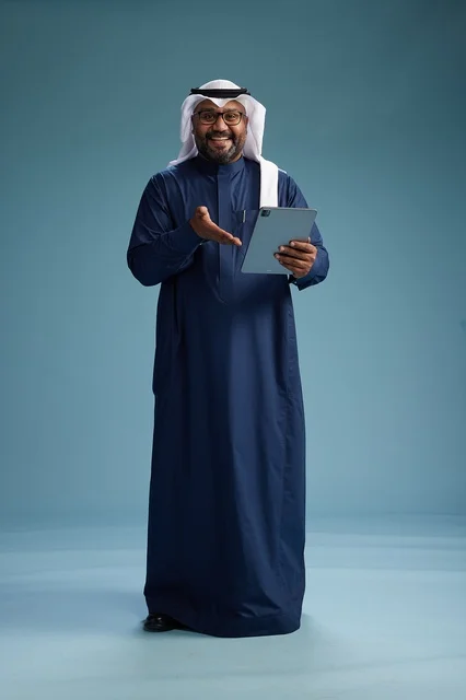 A Saudi man wearing a navy blue thobe and a white ghutrah is using a tablet, full-body shot, blue background.