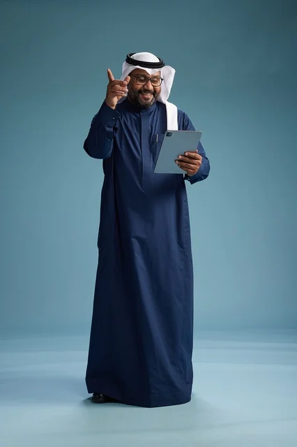 رجل سعودي يرتدي ثوب كحلي وغتره بيضاء يستخدم جهاز التابليت, الجسم بالطول الكامل, خلفيه زرقاء