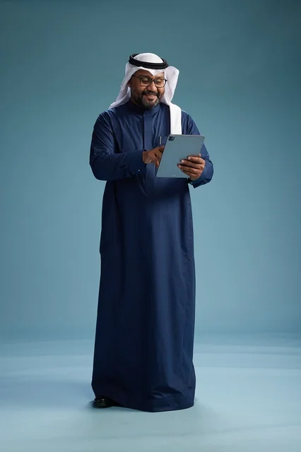 A Saudi man wearing a navy blue thobe and a white ghutrah is using a tablet, full-body shot, blue background.