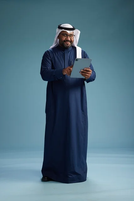 A Saudi man wearing a navy blue thobe and a white ghutrah is using a tablet, full-body shot, blue background.