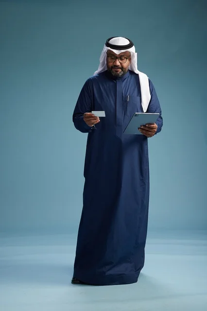 رجل سعودي يرتدي ثوب كحلي وغتره بيضاء يستخدم جهاز التابليت, الجسم بالطول الكامل, خلفيه زرقاء