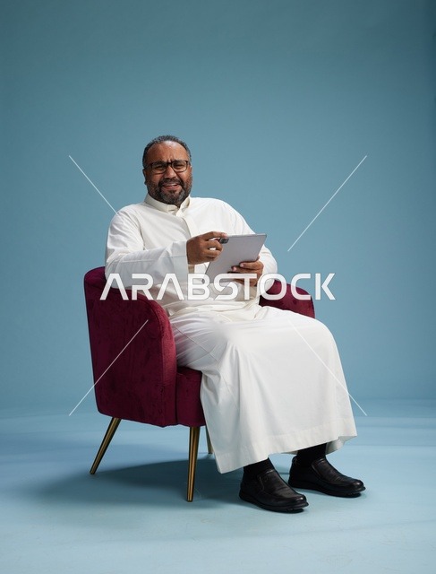 رجل سعودي بالزي التقليدي يحمل جهاز لوحي, استخدام التابليت المحمول, ايماءات وحركات متنوعه, خلفيه زرقاء