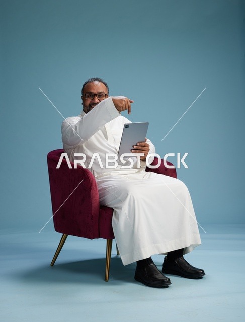 رجل سعودي بالزي التقليدي يحمل جهاز لوحي, استخدام التابليت المحمول, ايماءات وحركات متنوعه, خلفيه زرقاء