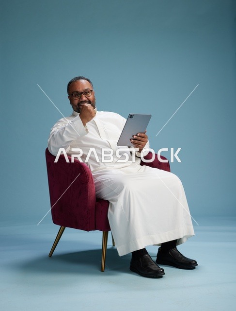 رجل سعودي بالزي التقليدي يحمل جهاز لوحي, استخدام التابليت المحمول, ايماءات وحركات متنوعه, خلفيه زرقاء