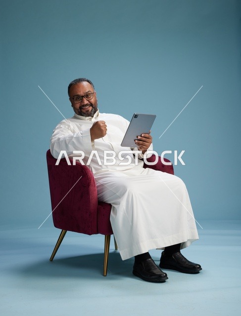 رجل سعودي بالزي التقليدي يحمل جهاز لوحي, استخدام التابليت المحمول, ايماءات وحركات متنوعه, خلفيه زرقاء