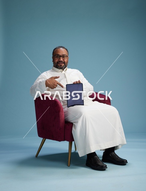 رجل سعودي بالزي التقليدي يحمل جهاز لوحي, استخدام التابليت المحمول, ايماءات وحركات متنوعه, خلفيه زرقاء