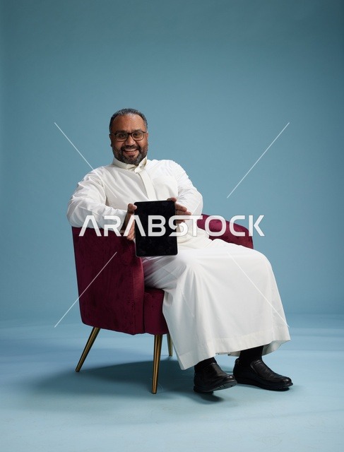 رجل سعودي بالزي التقليدي يحمل جهاز لوحي, استخدام التابليت المحمول, ايماءات وحركات متنوعه, خلفيه زرقاء