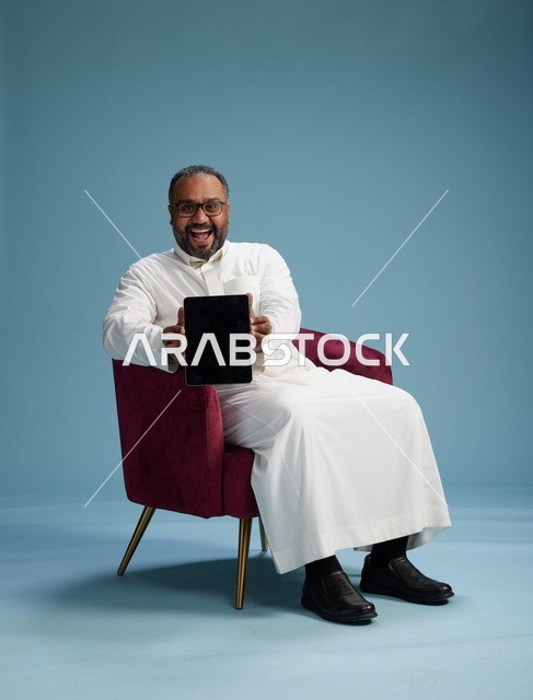رجل سعودي بالزي التقليدي يحمل جهاز لوحي, استخدام التابليت المحمول, ايماءات وحركات متنوعه, خلفيه زرقاء