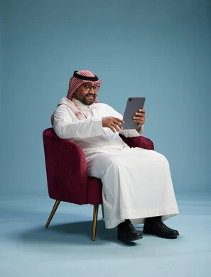 بورتريه لرجل سعودي  يرتدي الزي التقليدي  يجلس على كرسي احمر مريح يستخدم جهاز لوحي, خلفيه زرقاء