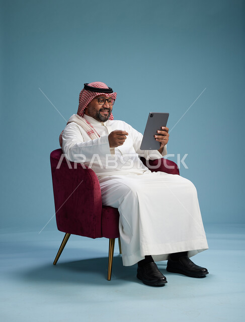 بورتريه لرجل سعودي  يرتدي الزي التقليدي  يجلس على كرسي احمر مريح يستخدم جهاز لوحي, خلفيه زرقاء
