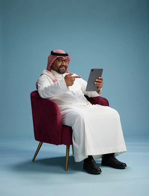 A portrait of a Saudi man wearing traditional attire sitting on a comfortable red chair using a tablet, with a blue background.