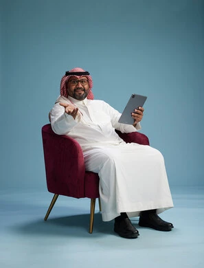 A portrait of a Saudi man wearing traditional attire sitting on a comfortable red chair using a tablet, with a blue background.