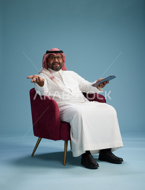 بورتريه لرجل سعودي  يرتدي الزي التقليدي  يجلس على كرسي احمر مريح يستخدم جهاز لوحي, خلفيه زرقاء