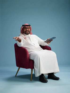 A portrait of a Saudi man wearing traditional attire sitting on a comfortable red chair using a tablet, with a blue background.