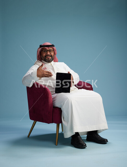بورتريه لرجل سعودي  يرتدي الزي التقليدي  يجلس على كرسي احمر مريح يستخدم جهاز لوحي, خلفيه زرقاء
