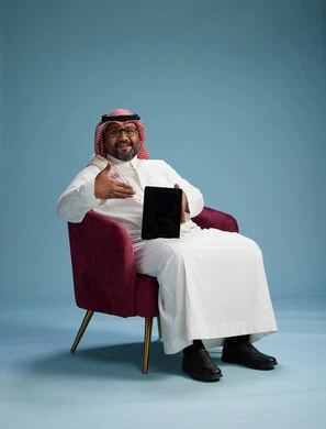 بورتريه لرجل سعودي  يرتدي الزي التقليدي  يجلس على كرسي احمر مريح يستخدم جهاز لوحي, خلفيه زرقاء