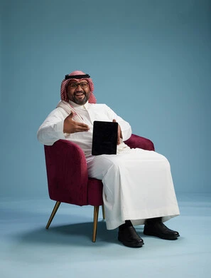 A portrait of a Saudi man wearing traditional attire sitting on a comfortable red chair using a tablet, with a blue background.