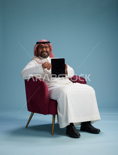 بورتريه لرجل سعودي  يرتدي الزي التقليدي  يجلس على كرسي احمر مريح يستخدم جهاز لوحي, خلفيه زرقاء