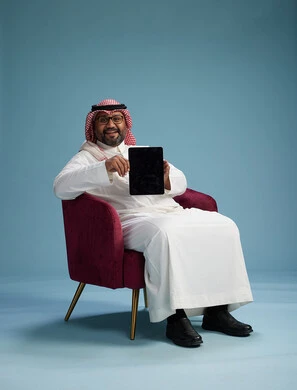 A portrait of a Saudi man wearing traditional attire sitting on a comfortable red chair using a tablet, with a blue background.