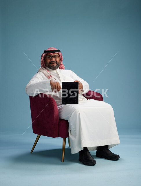 بورتريه لرجل سعودي  يرتدي الزي التقليدي  يجلس على كرسي احمر مريح يستخدم جهاز لوحي, خلفيه زرقاء