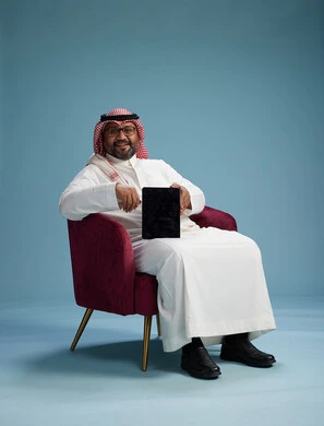 بورتريه لرجل سعودي  يرتدي الزي التقليدي  يجلس على كرسي احمر مريح يستخدم جهاز لوحي, خلفيه زرقاء