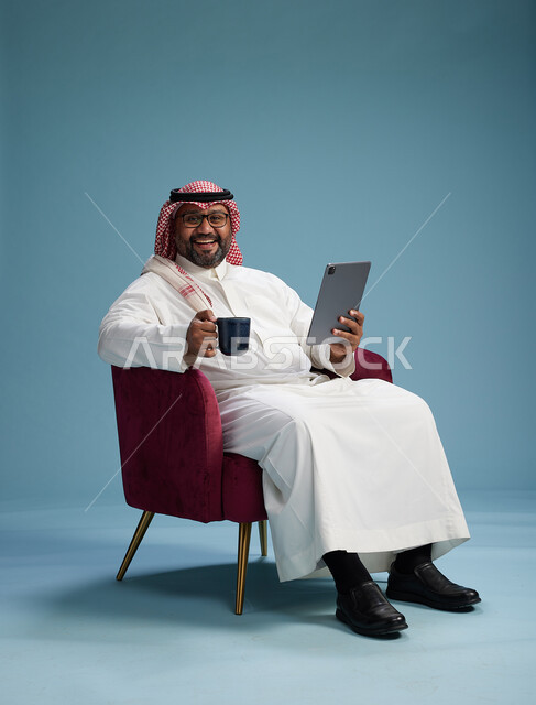 بورتريه لرجل سعودي  يرتدي الزي التقليدي  يجلس على كرسي احمر مريح يستخدم جهاز لوحي, خلفيه زرقاء