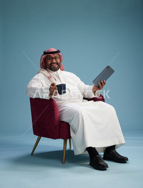 بورتريه لرجل سعودي  يرتدي الزي التقليدي  يجلس على كرسي احمر مريح يستخدم جهاز لوحي, خلفيه زرقاء