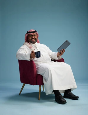 A portrait of a Saudi man wearing traditional attire sitting on a comfortable red chair using a tablet, with a blue background.