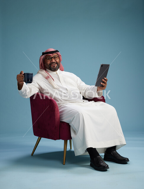 بورتريه لرجل سعودي  يرتدي الزي التقليدي  يجلس على كرسي احمر مريح يستخدم جهاز لوحي, خلفيه زرقاء