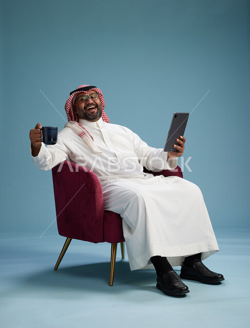 بورتريه لرجل سعودي  يرتدي الزي التقليدي  يجلس على كرسي احمر مريح يستخدم جهاز لوحي, خلفيه زرقاء