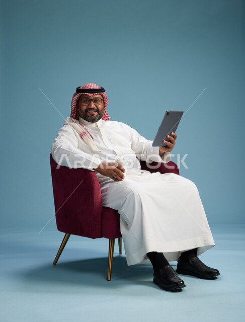 بورتريه لرجل سعودي  يرتدي الزي التقليدي  يجلس على كرسي احمر مريح يستخدم جهاز لوحي, خلفيه زرقاء