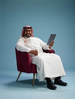A portrait of a Saudi man wearing traditional attire sitting on a comfortable red chair using a tablet, with a blue background.
