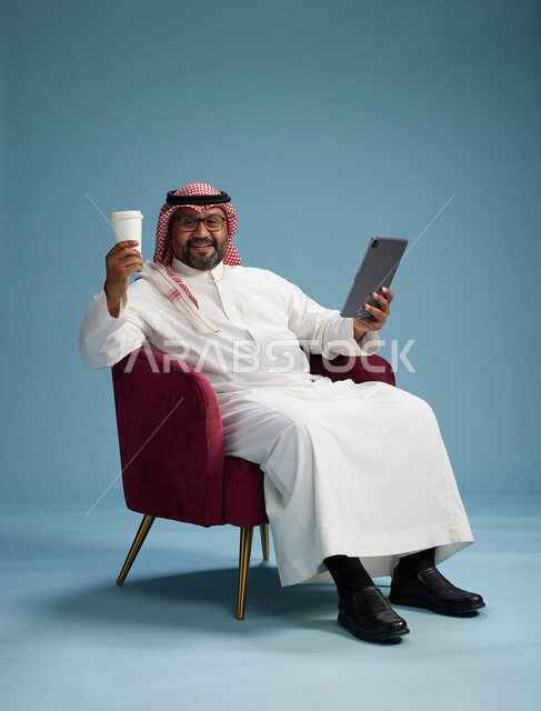 بورتريه لرجل سعودي  يرتدي الزي التقليدي  يجلس على كرسي احمر مريح يستخدم جهاز لوحي, خلفيه زرقاء