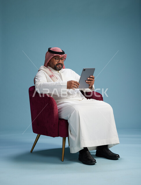 بورتريه لرجل سعودي  يرتدي الزي التقليدي  يجلس على كرسي احمر مريح يستخدم جهاز لوحي, خلفيه زرقاء