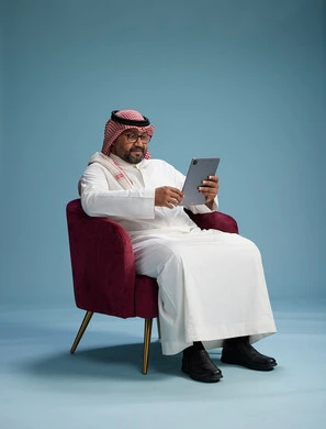 A portrait of a Saudi man wearing traditional attire sitting on a comfortable red chair using a tablet, with a blue background.