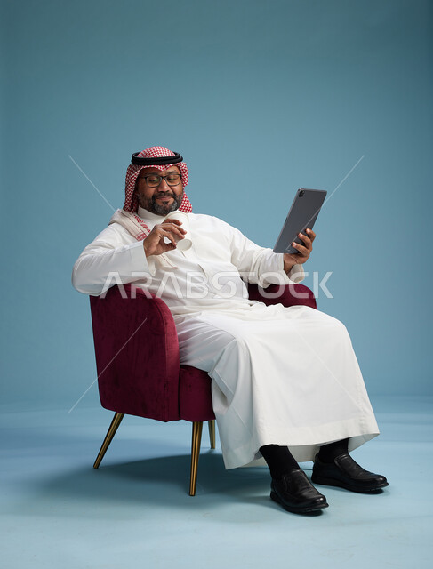 بورتريه لرجل سعودي  يرتدي الزي التقليدي  يجلس على كرسي احمر مريح يستخدم جهاز لوحي, خلفيه زرقاء