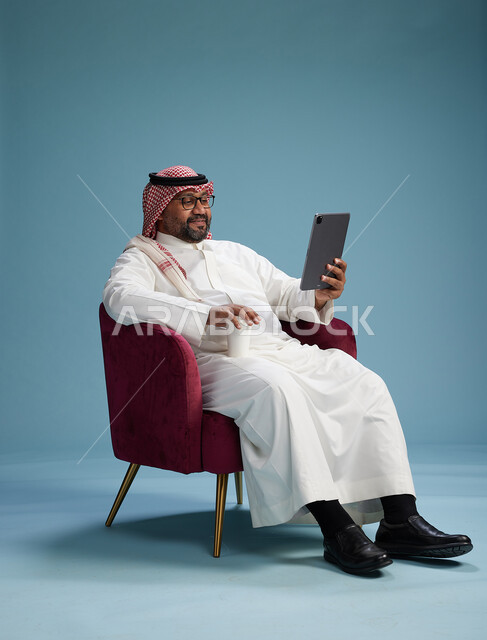 بورتريه لرجل سعودي  يرتدي الزي التقليدي  يجلس على كرسي احمر مريح يستخدم جهاز لوحي, خلفيه زرقاء