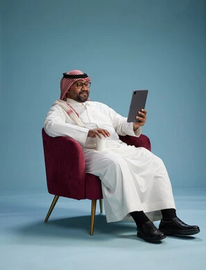 A portrait of a Saudi man wearing traditional attire sitting on a comfortable red chair using a tablet, with a blue background.