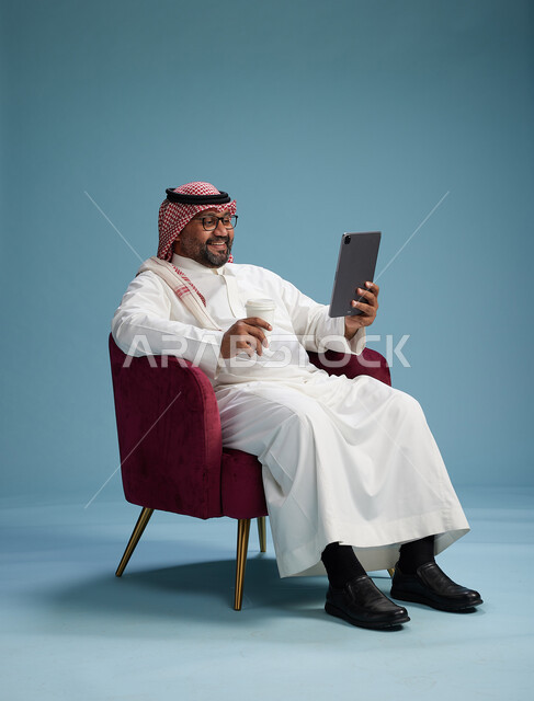 بورتريه لرجل سعودي  يرتدي الزي التقليدي  يجلس على كرسي احمر مريح يستخدم جهاز لوحي, خلفيه زرقاء