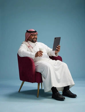 A portrait of a Saudi man wearing traditional attire sitting on a comfortable red chair using a tablet, with a blue background.