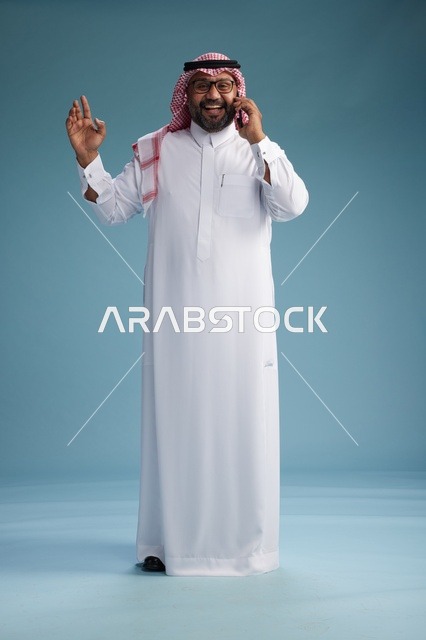 بورتريه لرجل سعودي يرتدي الثوب الابيض والشماغ يتحدث بالهاتف المحمول بإيماءة تعبيرية , الجسم بالكامل, خلفيه زرقاء